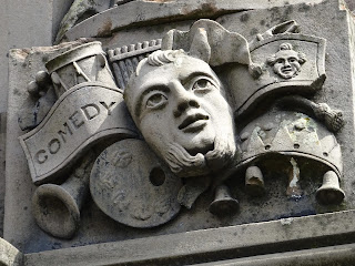 Detail from tomb of John Henry Alexander.  A photo of a carving of a face and musical instruments with a banner stating 'comedy' at the side.  Photo by Kevin Nosferatu for The Skulferatu Project.
