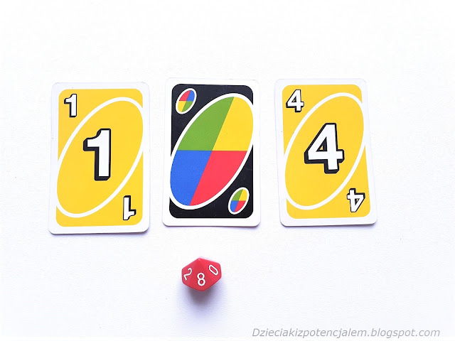 na zdjęciu karty uno o wartościach jeden i cztery a pomiędzy nimi leży karta joker, pod nimi kostka dziesięciościenna