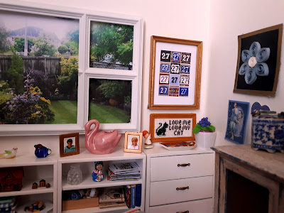 One twelfth scale modern miniature white room with a window overlooking a backyard. Under the window is a row of white bookcases and drawers containing various magazines, books, ornaments and craft supplies. On the wall are various handcrafted pictures.