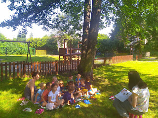 Grupa dzieci siedzi na kocu, który rozłożony jest na trawie. Dzieci słuchają pani bibliotekarki, która czyta im bajkę.