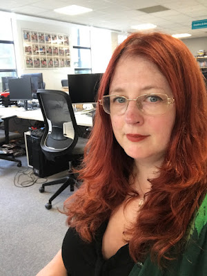 A redheaded woman in a mostly-empty office. She is looking into the camera.