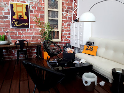 One twelfth scale modern miniature loft scene with brick exposed walls and wooden floors. At the back is a trestle desk and to the right is a seating area with a futon sofa, trolley coffee table and outdoor chairs. On the coffee table is an open storage box and a selection of papers, photos and postcards. Next to it is a small rubbish bin.