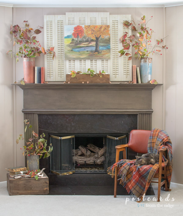brown fireplace mantel with shutters, old books, leaves, mid century chair with plaid blanket