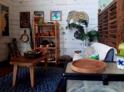 One twelfth scale modern miniature holiday house lounge with white shiplap walls and wooden floors. The decor is leaning towards vintage boho, woth a selection of vintage vases on a wooden bookcase, a guitar and ukulele propped in the corner and a selection of art pieces on the wall.