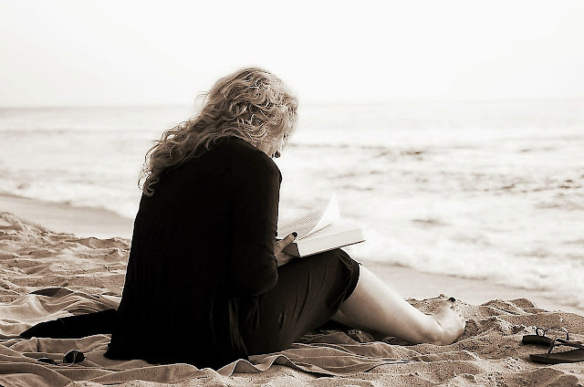 Una mujer de espaldas, leyendo un libro en la arena.