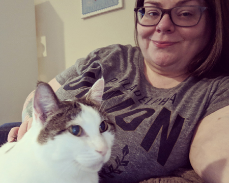 image of me sitting in a chair wearing a grey t-shirt and glasses, with my hair down; Olivia the White Farm Cat is sitting on my lap and I'm petting her