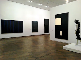 Interior view of an exhibition of black art, with several black work on display to the left, and a black Issey Miyake dress on display on a  mannequin on the right. 