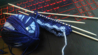 A cuff knit in 2 by 2 ribbing in self-striping yarn on double-pointed needles.  A hand-balled yarn ball is beside the needles. 