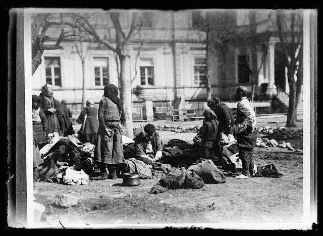 No Bath, No Food is American Rule in Balkans. A group of Balkan refugees in the yard of the American Red Cross Hospital at Monastir. To prevent the spread of Typhus and other diseases the Americans require every one asking aid of them to take a bath, which is provided in the building in the background. The food cards which they receive from the Red Cross unless the bath mark upon them has been punched. This particular group has just made its way back from an internment camp in Bulgaria. The man at the right in his bare feet has just had a bath, while the others are waiting to be called. During the bath their clothes are sterilized. The copper pot on the ground is a precious possession. It has been with this group throughout their four years of war travel