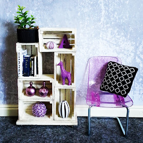 One-twelfth scale modern miniature scene containing a white shelving unit made of crates and a plastic glittery purple chair next to it. The shelving contains various decorative items in purple, white and black and the chair holds a black and white stitched cushion.