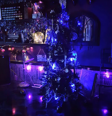 Christmas decorations at night The small black Christmas-tree is just right of centre in the picture. The camera looks diagonally towards the shelves. There is a Gothic arch mirror behind the Christmas tree. There is also a lamp, turned off, in the shape of a book, with a Harry Potter themed cover that says Liber Lux behind the tree. The tree is on a shiny simulated marble worktop. A computer screen is just visible on the far left, but it is turned off. There is a grey vintage-style desk-lamp underneath the shelves. The shelves are black with ornate Victorian brackets. Hanging from the lowest shelf is a string of lantern-shaped string lights, quite small - the panes are green and purple. On the first shelf there are ornate boxed of art materials and a glass skull filled with small white LED lights. The second shelf is farlanded with iridescent tinsel that seems to be glimmering in the low light. On the second self there is a string of tiny purple bat-shaped lights reaching upwards and a rack with coloured markers. The small black Christmas tree has bat-shaped lights that seme very blue in the photograph. There is a row of round purple lights running under some black dado rail; it illuminates grey wallpaper depicting Gothic arches so it loos purple, and reflects off the glossy worktop.The image is dark, it is taken at night to maximise the effect of the string-lights. The wall is visible in the upper right and it is a very dark purple. There is a Highlands and Islands region sticker for the Scottish Green Party on the Gothic mirror behind the Christmas tree.