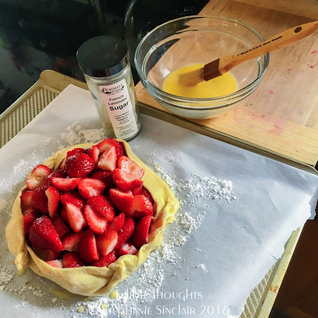 Image of a baking sheet lined with parchment paper dusted with powdered sugar. On top is a puff pastry filled with a pile of cut up strawberries. A glass jar next to it is labeled as "French Lavender Sugar." On a nearby bamboo cutting board is a glass bowl with a brush and whisked egg.