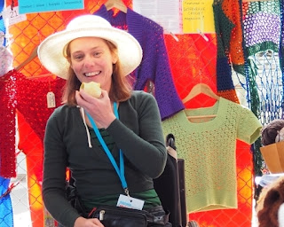 Jodie is seated, smiling with an apple in her hand. Behind her are garments hanging on the fence. From left to right, red beaded crochet lace top and belt, purple lace stitch cardigan, green 'petal pullover' short sleeved crocheted top, 'Woodstock duster jacket' in blue, green, orange and purple crochet mesh. 