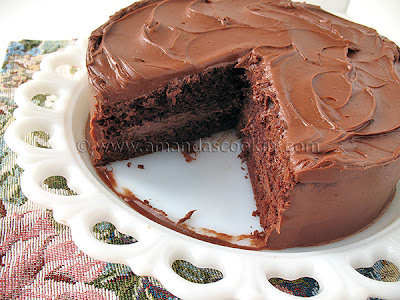 A close up photo of a Nigella\'s old fashioned chocolate cake resting on a white cake stand with a slice removed.