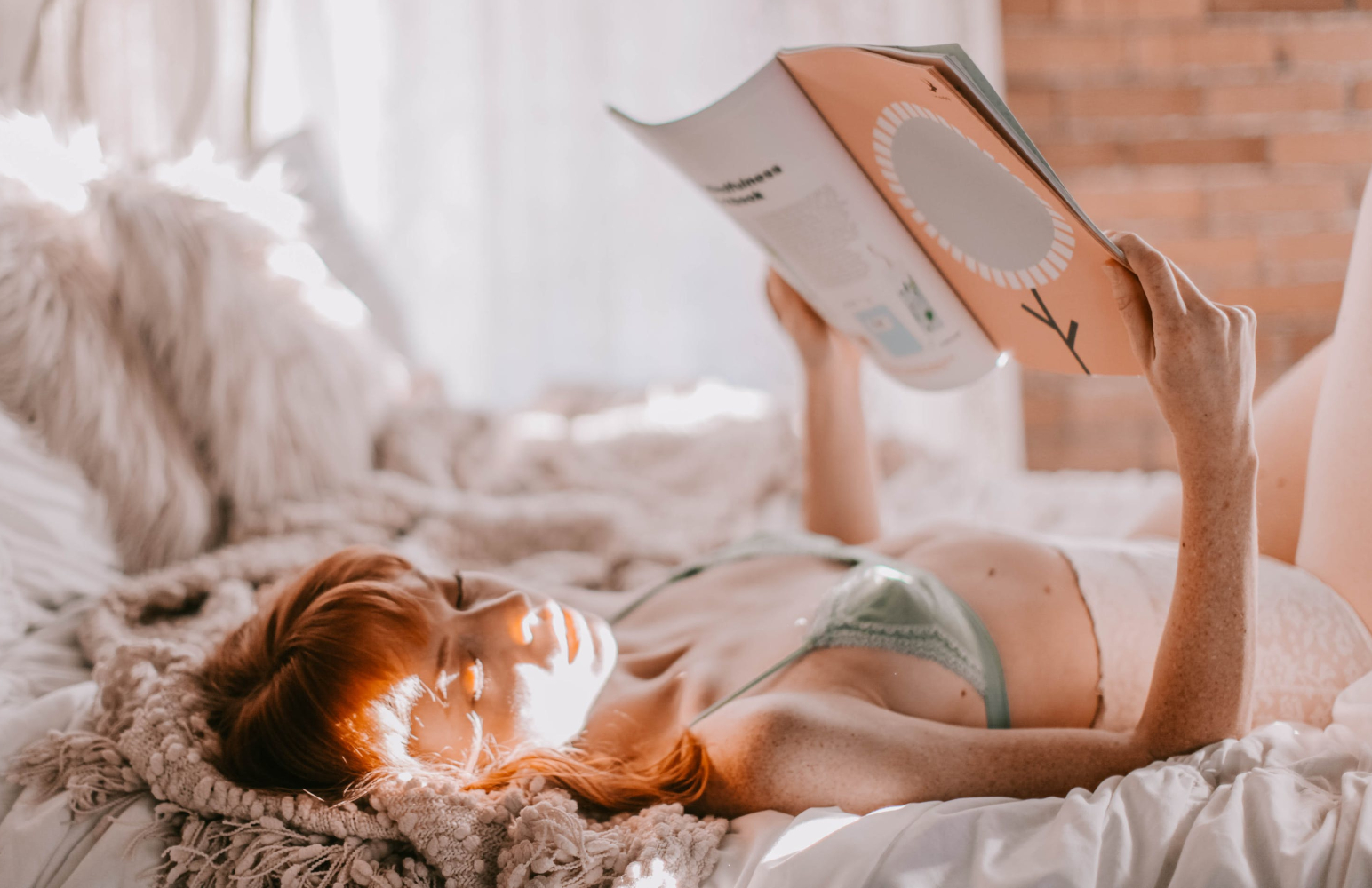 ginger woman in blue bralette is posing with magazine on a bed