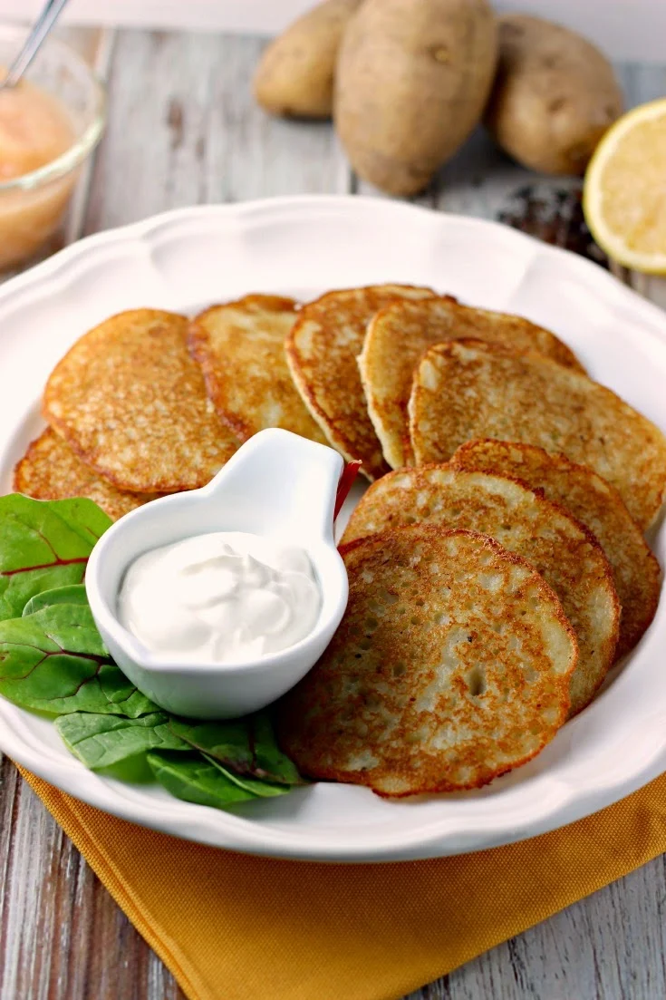 Easy Potato Pancakes by Renee's Kitchen Adventures on a white plate with a lettuce garnish and sour cream. Applesauce and potatoes in the upper top background.