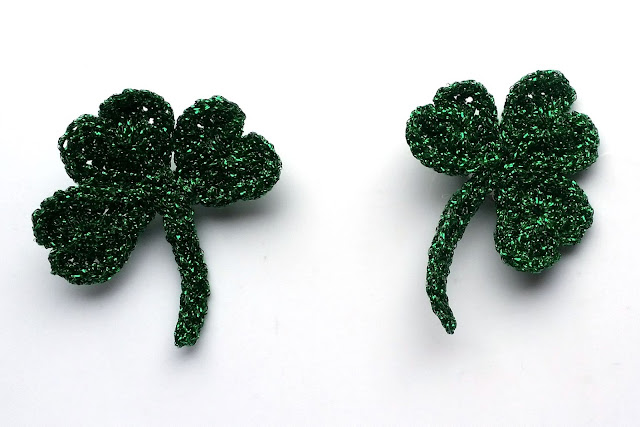 Two completed crocheted shamrocks made of green sparkly metallic yarn.  The shamrocks have three heart-shaped leaves, joined at their points and connected to a stem.