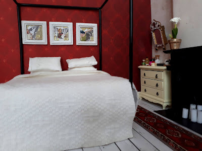 One-twelfth scale modern miniature bedroom with a black metal four-poster bed with white bed linen and a red wall behind with three framed art works above, On the right-hand wall is a fireplace with candles inside, and next to it a chest of drawers with various cosmetics on the top, and a enamelled mirror on the wall above.