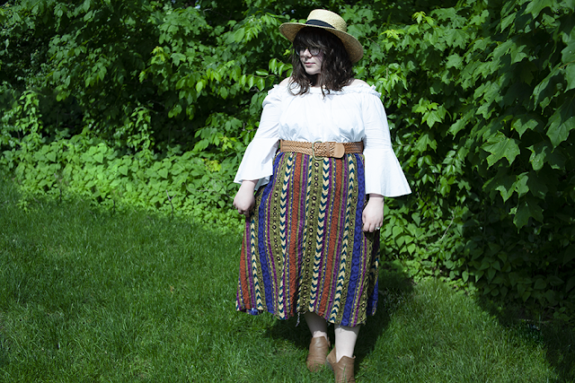 An outfit consisting of a large straw sunhat, white ruffle off the shoulder blouse, a printed maxi skirt, and brown booties.