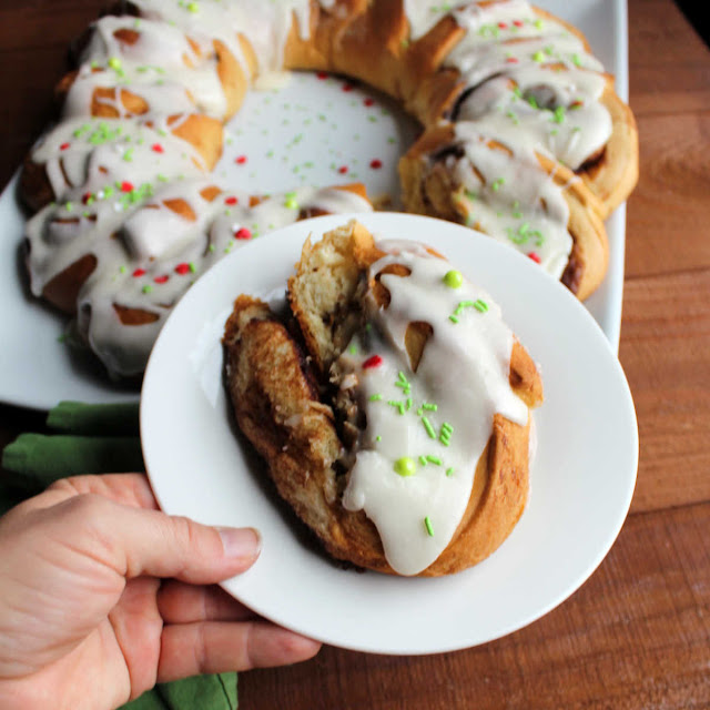 Soft and sweet cinnamon rolls take on a festive wreath shape. They are a perfect holiday breakfast or something fabulous for overnight guests. hand holding cinnamon roll on plate with remaining cinnamon roll wreath in background