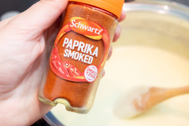 A close up of a hand holding a bottle of smoked paprika over a creamy sauce in a metallic silver pan.