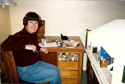 A woman in her twenties, sitting at a desk next to a table holding a one twelfth scale bay villa dolls' house. She has a paint brush in her hand and a miniature project on the desk.