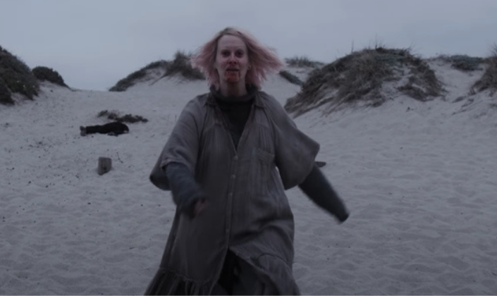 Imagem de capa: a personagem T.B. Karen, interpretada por Sarah Paulson, em roupas velhas e cinzentas e com os cabelos loiros sujos, sangue sujando sua boca e queixo, andando confiante por uma praia fria com céu cinzento ao fundo.