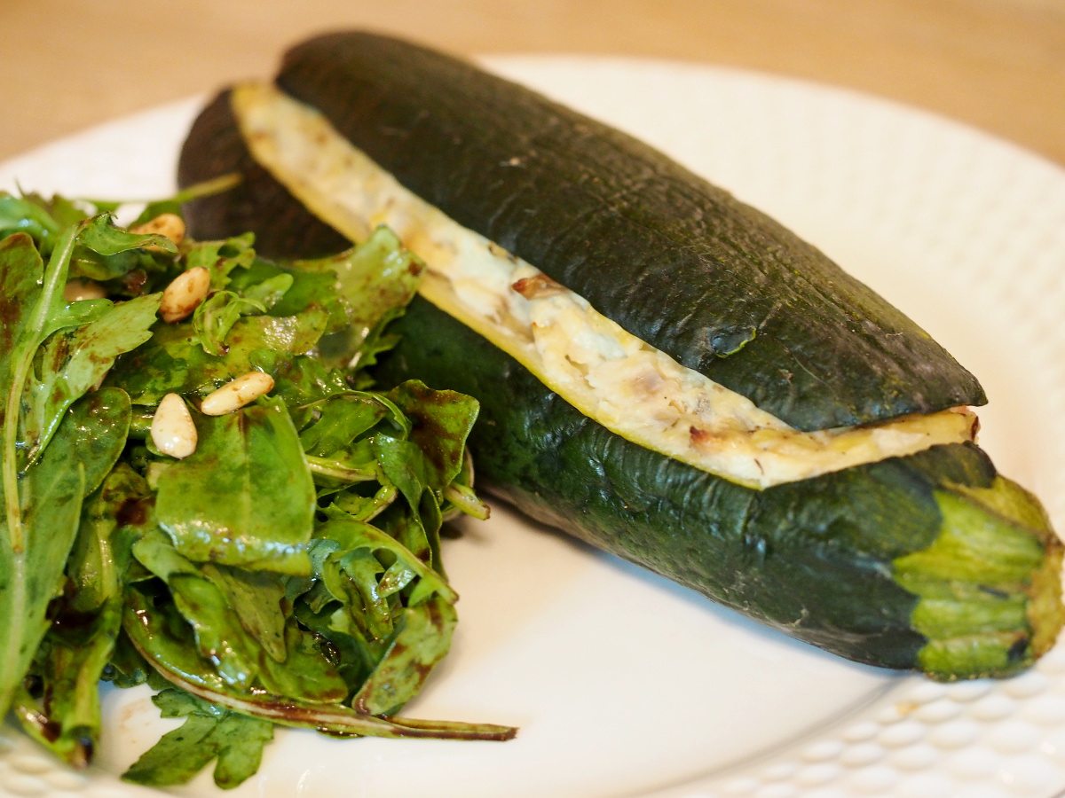 Eine gefüllte Zucchini auf einem Teller, daneben liegt etwas Rucolasalat mit Pinienkernen.