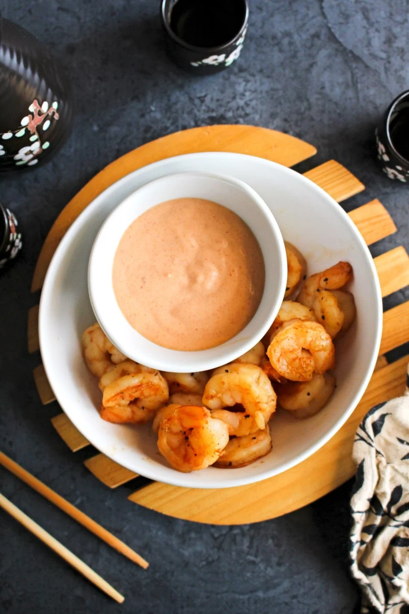 Top view of yum yum sauce in a white bowl sitting in a larger white bowl filled with cooked shrimp.  Bowl is on a wood rack sitting on a grey background.