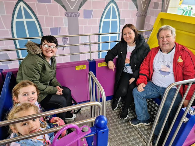 Image of a family inside a carriage on the Queen's carriage ride at Peppa Pig World. The carriage is large and has benches at either end. It is decorated in regal colours and is bold and bright. It runs along a high wire track around the park. There are 3 adults in the carriage and 2 children.