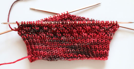Detail of the beginning of a hand-knit sock on the needles on a white background.