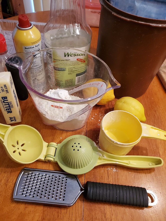This is all the ingredients in a OXO black handled mixing bowl for lemon copycat Starbucks loaf cake. There is a stick of butter, lemon, measuring cup, real lemons, flour, lemon juicer in the photo.