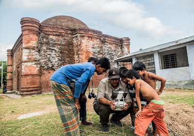 Niños observan la pantalla de una tablet, conectada a un mando de dron, mientras la sostiene un hombre. Detrás, la Mezquita de las Nueve Cúpulas, en Bangladesh.