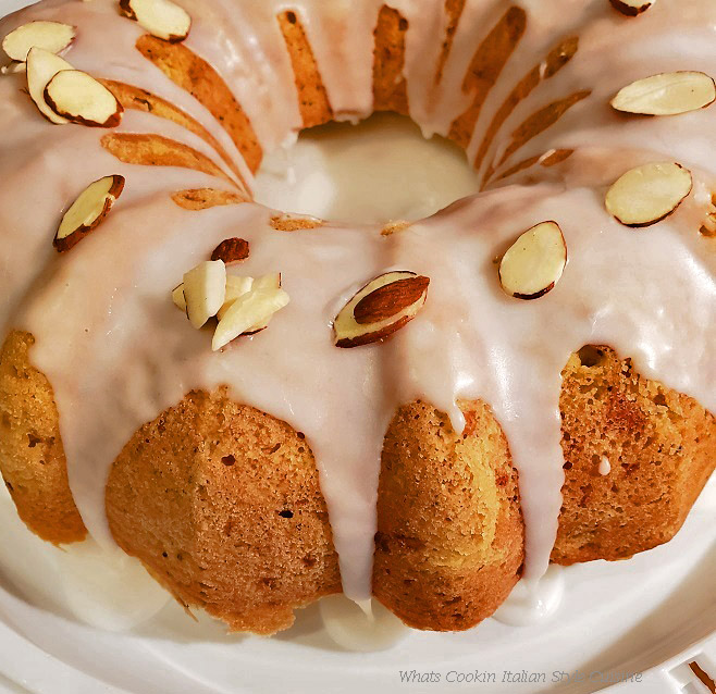 This is a bundt cake frosted with white frosting and sprinkled with toasted almonds. The cake is a zucchini almond and poppy seed flavored cake and sitting on a white cake taker that is plastic.