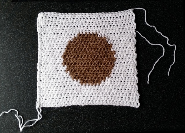 A crocheted white square with a tan intarsia circle in the middle, resting on a black background.  White loose ends can be seen around the edges. This is the right side of the square.  The left hand edge is straight like the top and bottom edge, while the right hand edge is 'bumpy' because it is made of raw row ends.