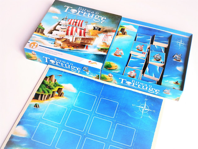 na zdjęciu pudełko od gry ora rozłożona plansza przedstawiająca morze a na nim dwanaście kwadratów oznaczających pola startowe dla okrętów