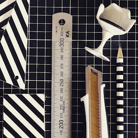 Flat lay of a pencil, utility knife, metal ruler and a one-twelfth scale modern miniature tulip chair along with a rectangle of black and white striped wood, plus some offcuts.