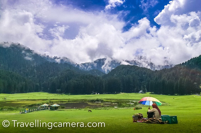 A visit in an year to Khajjiar  has made me click less photographs and rather experience different weather there. I was in Khajjiar during monsoons and this is probably the best season to experience lush green landscapes all around. Khajjiar, at times, is not accessible during winters because roads get closed due to snowfallThis is the first view of Khajjiar lake when you are coming from Dalhousie. This is the point where you should start looking for parking slot. Road condition is usually not very good during monsoons. Maintaining roads in hills is very challenging, but still Himachal has been aggressive in maintaining it's roads. Especially roads in tourist regions.During Monsoons, it's very important to keep umbrella with you, even if sun is shining and there is no patch of clouds. This change in minutes and hence very unpredictable. Our day started with clear sky and by the time we reached Khajjiar it started raining heavily. Fortunately we were carrying few umbrellas and borrowed one from our cab driver.While it was raining, we took a table in one of the restaurants around Khajjiar Lake. It was lunch time and we thought of utilizing this time and had lunch. I am forgetting the name of the place where we had lunch and it was good Punjabi food.A few kilometers ahead there is a temple with this huge Shiva idol. Now let me share a very useful tip. If you came to Khajjiar from Dalhousie in a bus and want to go back in bus only. Reach this place 30 minutes back. Usually there are lot of folks who board bus in Khajjiar and it becomes very challenging to get in. Since this temple is a stop before Khajjiar, there is good probability to get in comfortably and also find a seat. There are 2 buses from Khajjiar to Dalhousie between 1pm to 3pm.So if you come early in the morning, the timings would suit you.These days paragliding  is also quite popular activity around Khajjiar. One flies from a surrounding hill. You need to climb of the hill, so be sure about it. After flight, depending upon the flow of wind you can have a top view of Khajiar, but landing happens in village behind the shiva temple. I haven't done paragliding here so don't know the charges. Bir Billing is best place for Paragliding in Himachal Pradesh and I had that experience.Another view of Shiva from road connecting Khajjiar with ChambaMonsoon is a good time to taste local produce. Local cucumber is much tastier than what we get in cities through cold storage. During Monsoons, you would see lot of folks selling fresh fruit salad around Khajjiar and what can be best snack when travelling in himalayas.Clouds make this place more beautiful. I made some videos of clouds covering this place and going upwards to play in woods, but unfortunately we lost those videos due to card corruption. As I see some herbs vendor in above photograph, so let me warn you about a group of folks who sell you local herbs (don't know what that means and I never bought). But I have heard bad experiences of folks who dealt with these vendors. Please note that most of them are not local folks. and things they sell are not locally produced. They bring stuff from Jammu or Pathankot and sell to tourists. So if you can judge the originality of stuff being sold, go have a talk. Otherwise avoid even talking.  Pre-wedding shoots and post wedding shoots are getting popular in our country. And I noticed lot of couples here at Khajjiar with professional photographers. Some of these photographers roam around the lake and click photographs to hand-over in form of prints. Some of the photographers had come with couples, were indulged in finding appropriate location and guiding these couples for some unique shots.I also wanted to do a shoot with my model, but this time she was acting like celebrities. And even weather was not favoring us. So this time, I couldn't click much.Here is path outside the green meadows, which is used for horse riding. You can hire a horse to have a round of Khajiar or roam around the neighboring villages. I never took the other route where these folks promise to show village and apple orchards.  Carefully notice this photograph. Imagine the view when these clouds are slowing coming out of these forests. This was the best moment and experience at Khajjiar.There are lot vendors selling toys for kids. Urvi also wanted few but settled at one, which didn't reach home safely :). We had long day, so that was expected... I like some of these things at tourist places which provide opportunities for local folks. Some selling these toys, few making fruit salad and others selling juicy bhuttas. And all these things are also available at reasonable costs if you compare with prices quoted in bigger cities like Delhi. And it's not about money, it's more about purity and love of these local folks.This was shot when I was just leaving for Chamba. These colorful baloons in front of these green landscapes looked awesome. Over the years I have seen this place transforming, in good as well as bad ways. Local authorities don't allow people to play any kind of sport on these lawns but there areNames of horses around Khajjiar are very interesting :) ... Romeo, Chetak etc..  If you intend to stay around Khajjiar, there are 3 reasonable options - HPTDC hotel, HPPWD Guest-House and Forest Guest-House. Apart from there are there are various private properties.