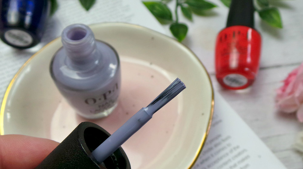 Image shows a close up of the nail polish brush of a lilac grey polish.  The open bottle is sat on a pink dish in the background