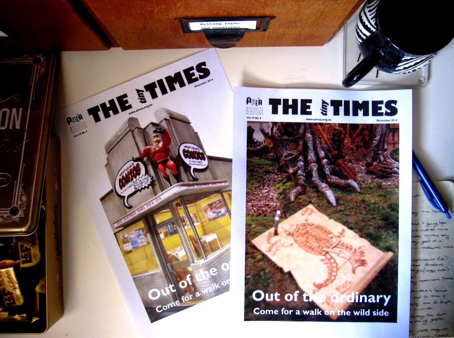Desk with a tin of chocolate bars, two test covers for The tiny Times, a notebook and pen and an empty tea mug on it.