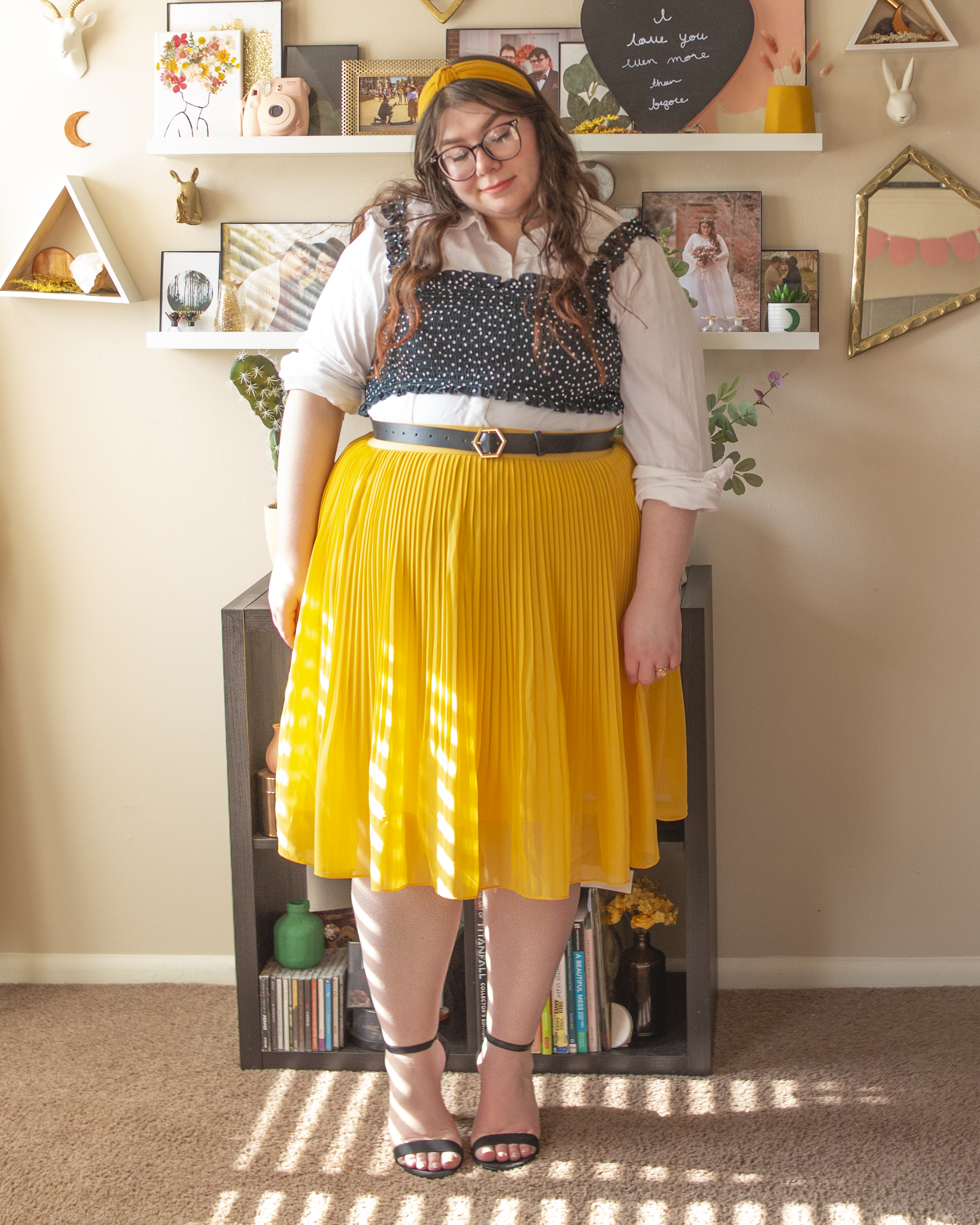 An outfit consisting of a white cotton long sleeve button up blouse with a black white white swiss dots crop top layered over and tucked into a yellow pleated midi skirt and black ankle strap heeled sandals.