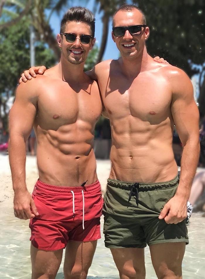 two-shirtless-fit-beefy-gay-young-bros-smiling-sun-tanning-beach-shorts-sunglasses-abs