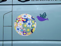 Photograph of the side of eggshell van. Dasies with colour elements; red, blue green, purple are arrange together and have been pulled out into a 3d ball shape, printed flat. A stylised dove in purple and green is flying next to the ball of flowers.