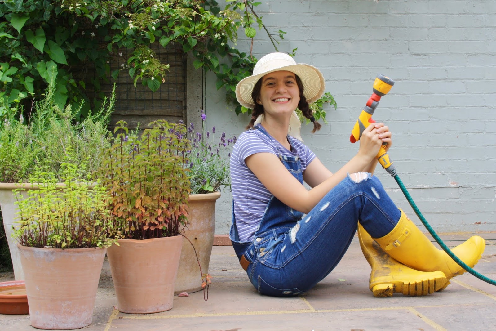 Abbey, wearing blue dungarees and a straw hat, sits among pot plants in a garden, holding a yellow hose