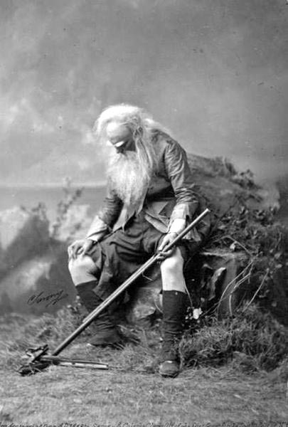black-and-white studio set-up of a very old man with a long beard seated in the countryside, with a gun barrel in his hand and broken pieces of a gun on the ground