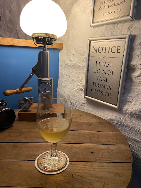 A pint of cider on a wooden table, in front of a lamp made out of a piece of machinery. The walls in the room are rough, whitewashed stone.