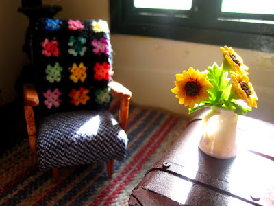 Modern dolls' house miniature armchair with afghan rug on the back, and chest with a vase of sunflowers on top.