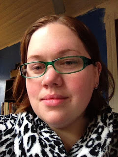 Author with wet auburn hair, rosy skin, green plastic glasses, no makeup, wearing a snow leopard print fuzzy bathrobe. Wall is medium blue in background with a bookshelf visible