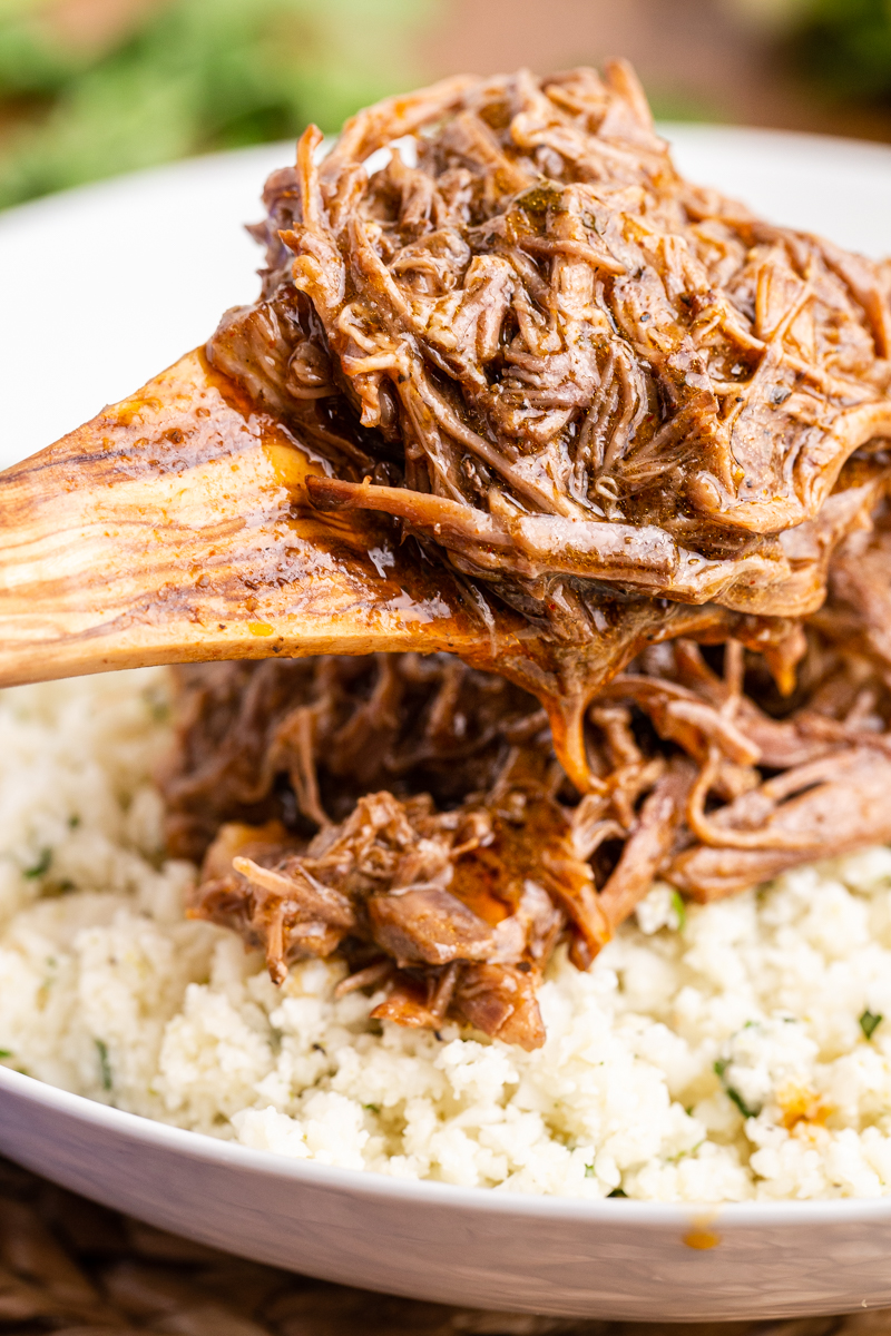 Closeup photo of someone spooning Instant Pot Mexican Shredded Beef  into a white bowl over cilantro lime cauliflower rice.
