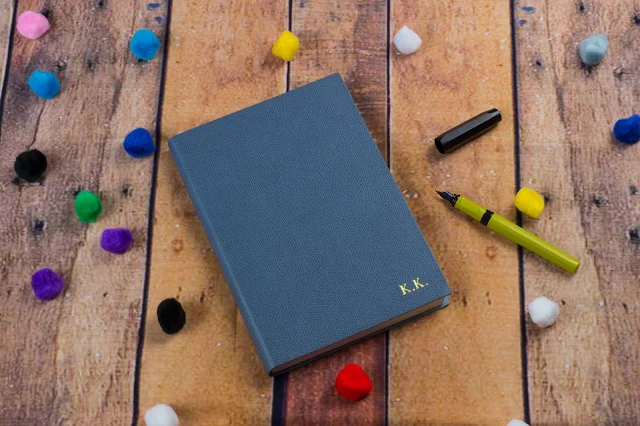A beautiful leather journal with gold embossed letters saying KK on floor boards next to an open fountain pen
